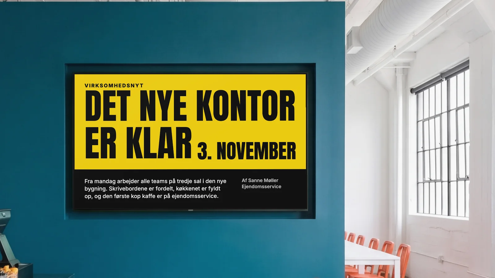 Personalekantine med en skærm indfældet i en dyb petroleumsblå væg, et glimt af disken og orange stole ved siden af. Skærmen viser Virksomhedsnyt: Det nye kontor er klar den 3. november.