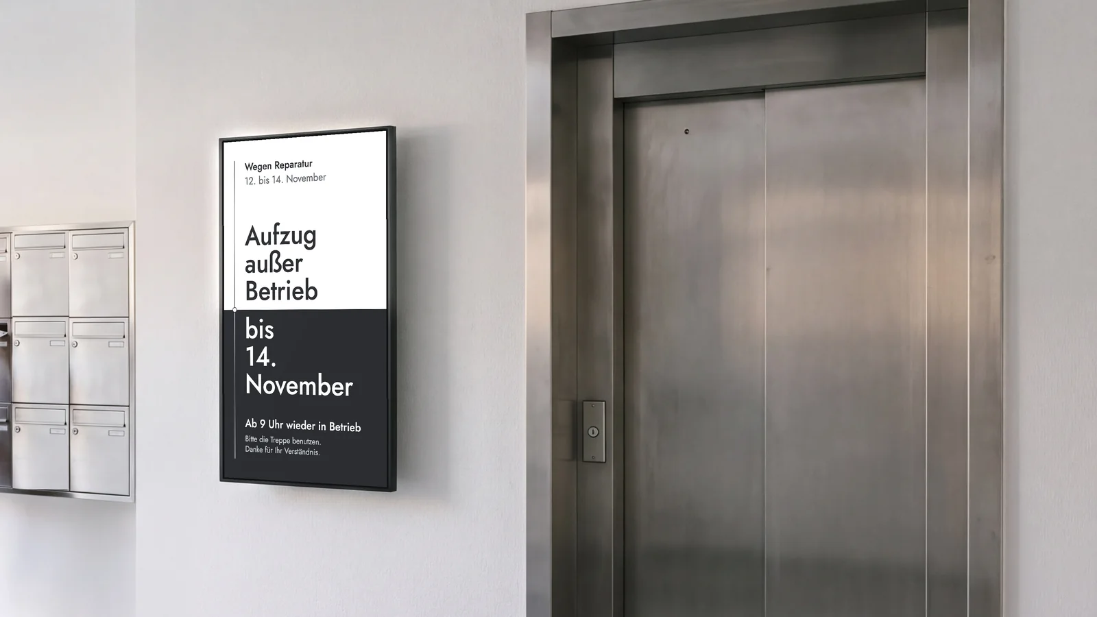 Eingangsbereich eines Mehrfamilienhauses mit einem Bildschirm im Hochformat an der weißen Wand zwischen Briefkästen aus Stahl und einer großen Aufzugstür aus Stahl. Er meldet: Aufzug außer Betrieb bis 14. November, Reparatur vom 12. bis 14. November, ab 9 Uhr wieder in Betrieb.
