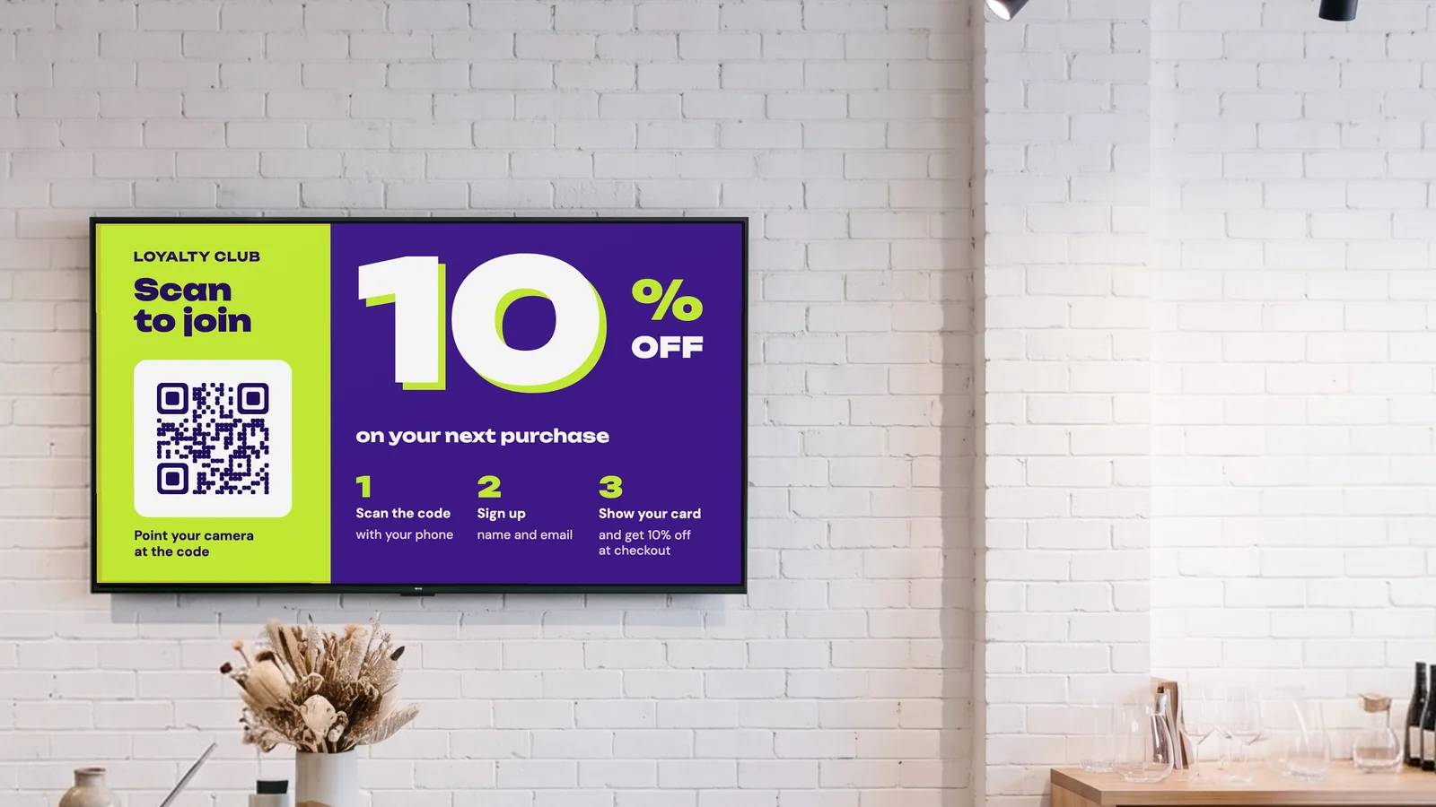 Gift and home goods shop with a screen on the white brick wall behind the till, dried flowers on a terrazzo counter and ceramics on shelves at the side. The screen promotes the loyalty club: scan to join and get 10% off your next purchase.