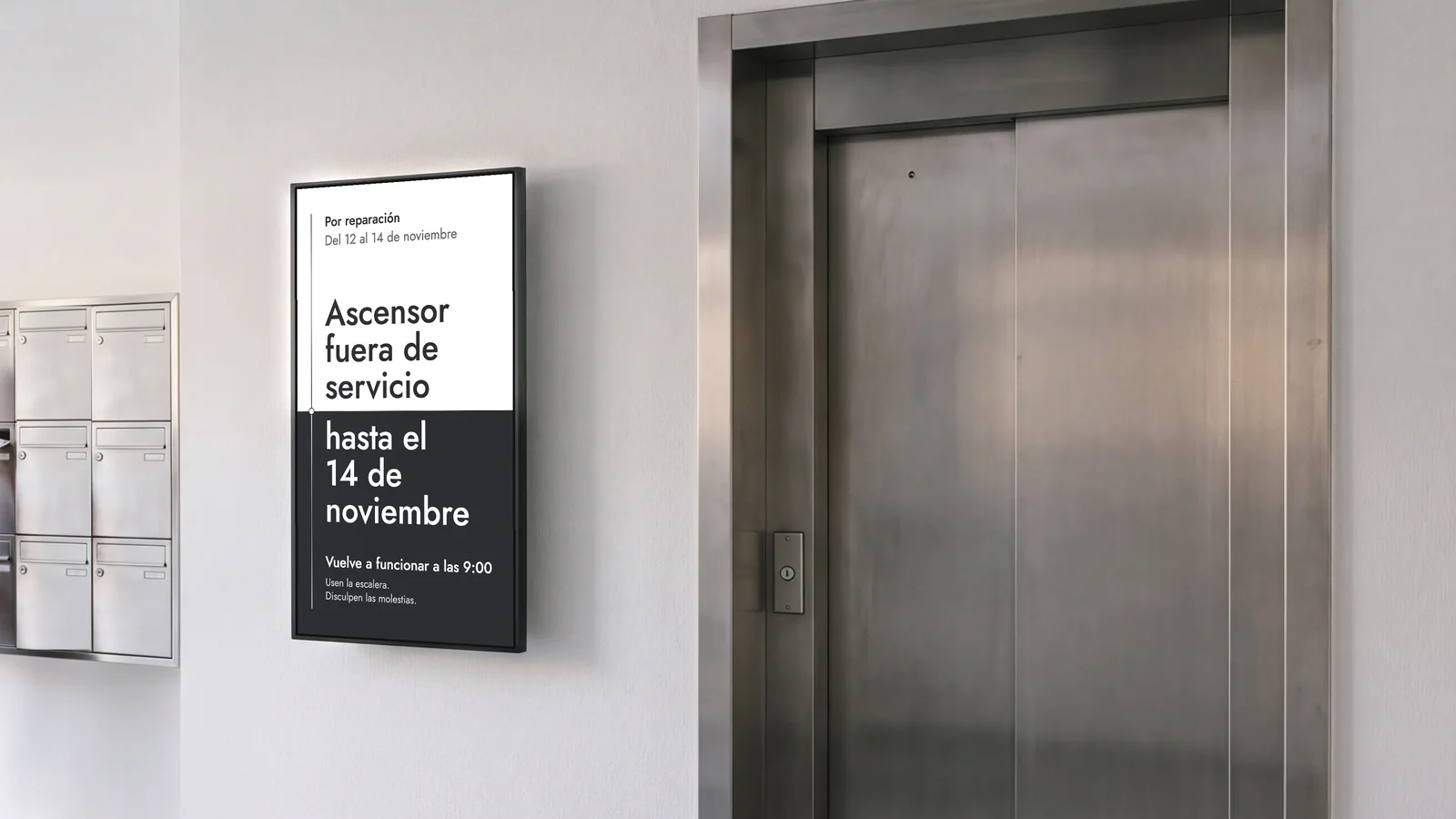 Portal de un edificio de viviendas con una pantalla en vertical en una pared blanca, entre buzones de acero y una gran puerta de ascensor de acero. La pantalla indica: ascensor fuera de servicio hasta el 14 de noviembre, reparación del 12 al 14, vuelve a funcionar a las 9:00.