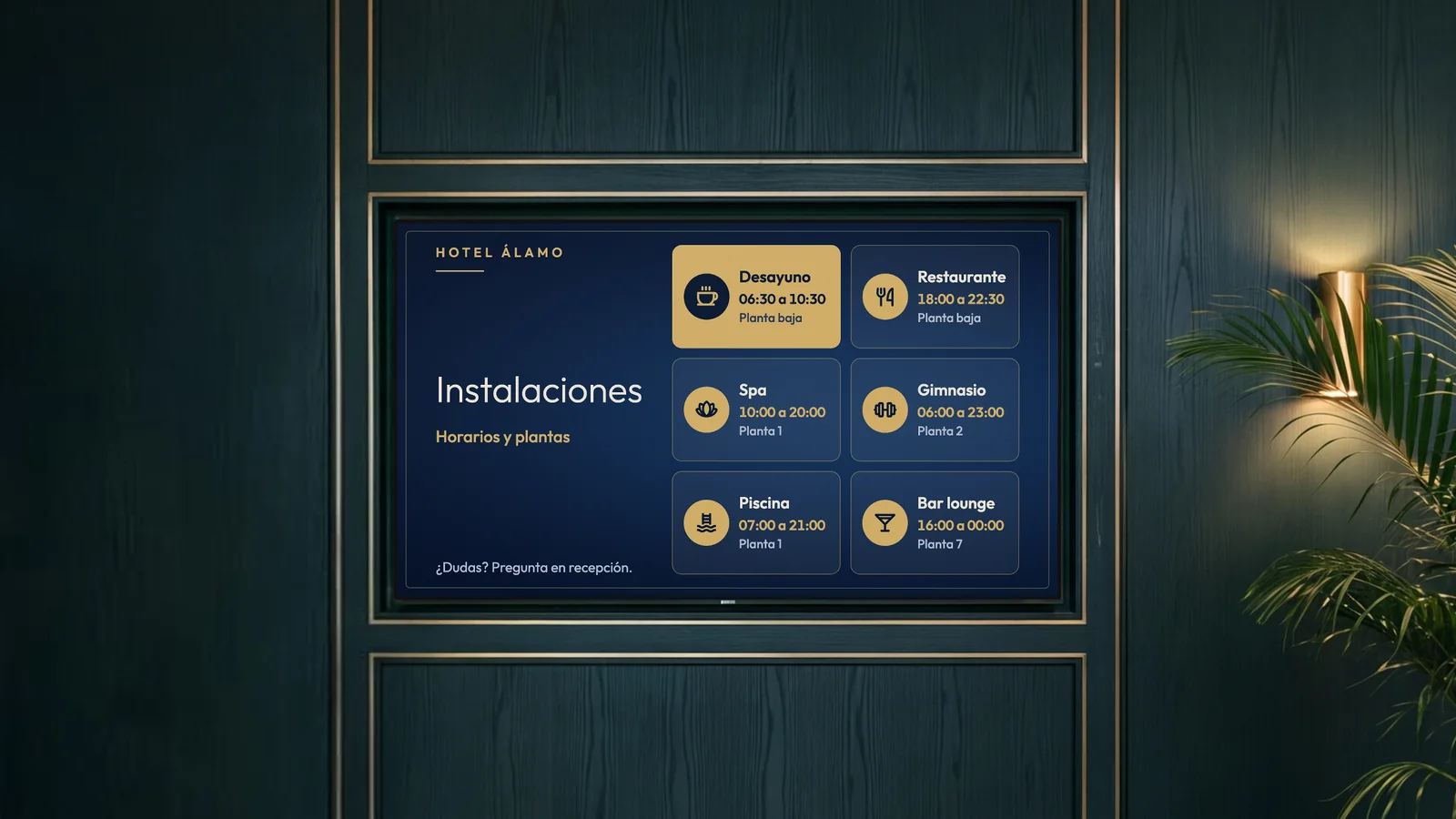 Vestíbulo de ascensores de un hotel urbano con paneles de madera verde oscuro, detalles de latón, un aplique dorado y una palmera junto a la pantalla. La pantalla muestra las instalaciones del Hotel Álamo: desayuno, restaurante, spa, gimnasio, piscina y bar lounge.