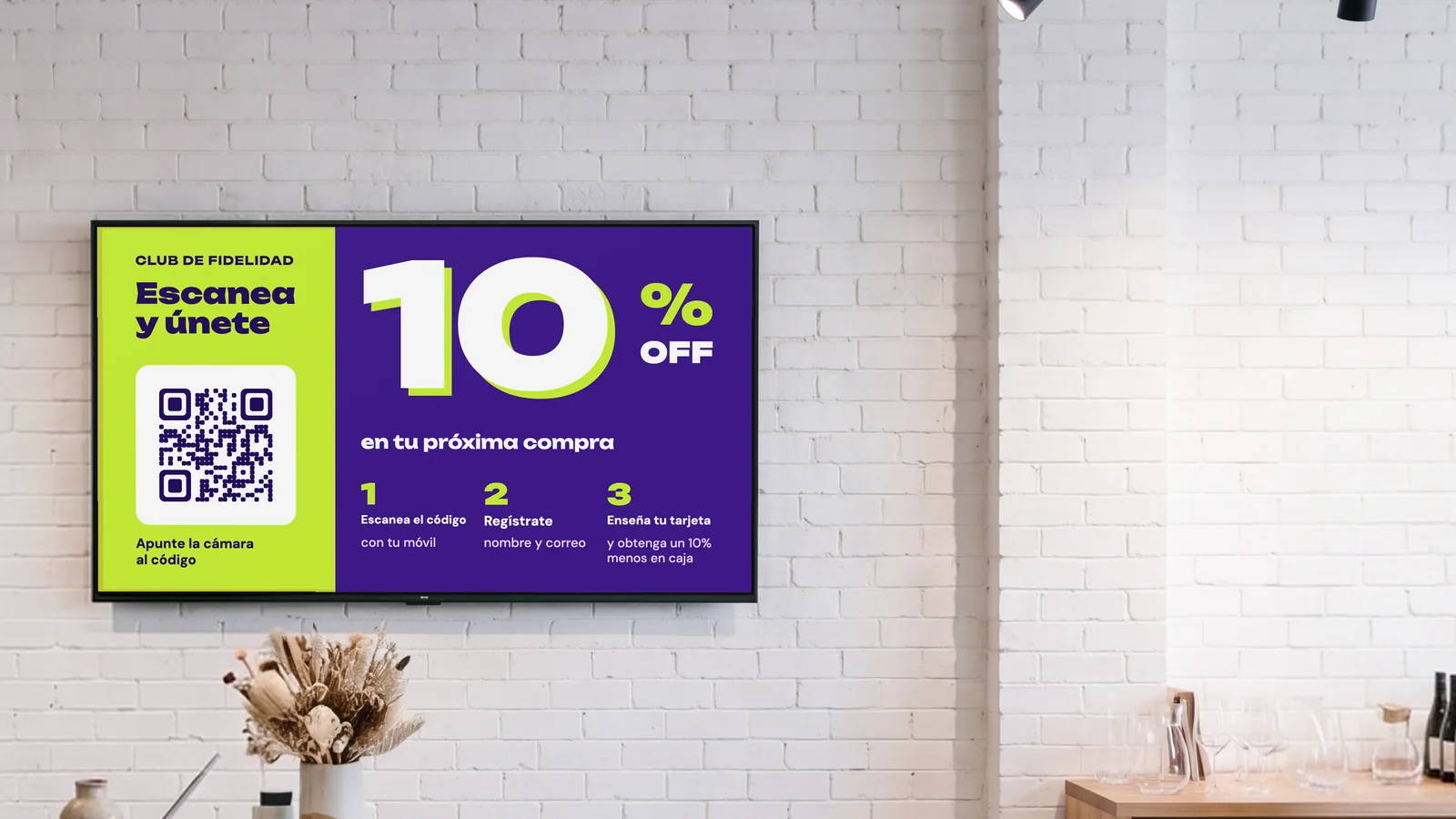 Tienda de regalos y decoración con una pantalla en la pared de ladrillo blanco detrás de la caja, flores secas sobre un mostrador de terrazo y cerámica en las estanterías del lateral. La pantalla anuncia el club de fidelidad: escanea y únete, 10 % de descuento en tu próxima compra.