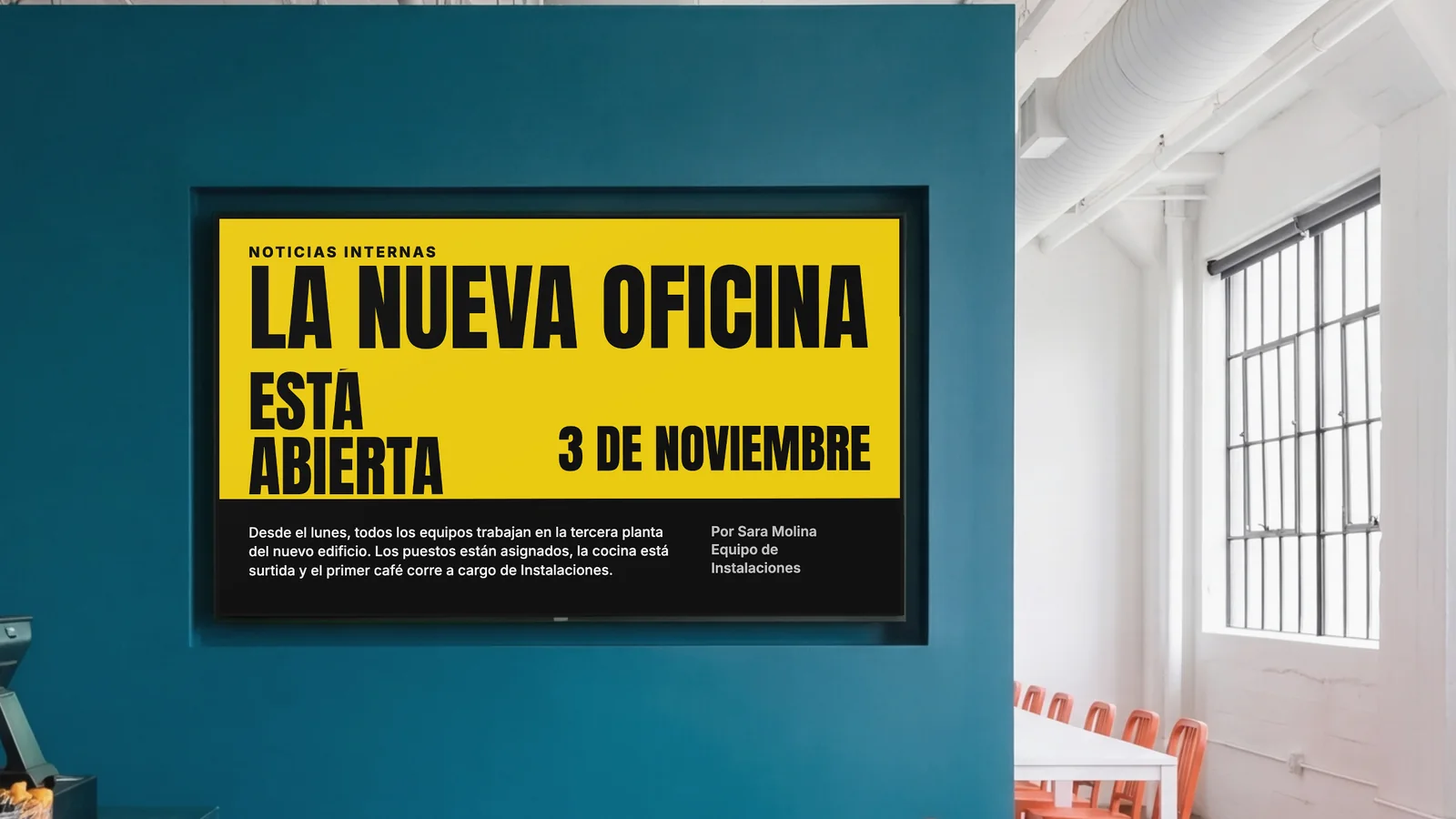 Comedor de empresa con una pantalla encajada en una pared verde azulado oscuro, un rincón del mostrador y sillas naranjas a los lados. La pantalla muestra Noticias internas: la nueva oficina está abierta, 3 de noviembre.