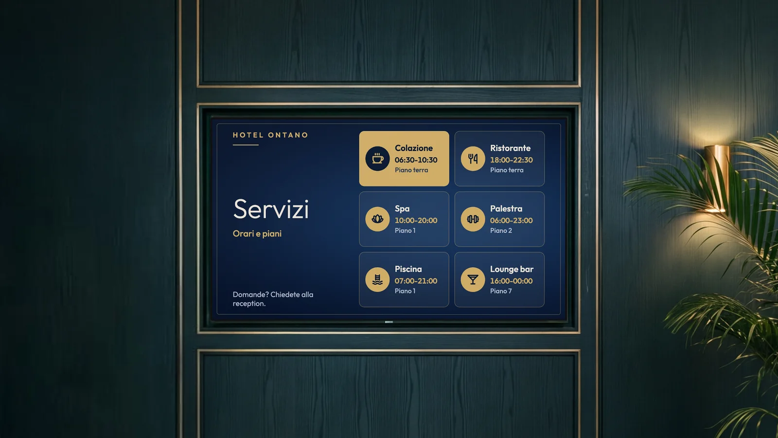 Zona ascensori di un hotel in città con boiserie verde scuro, dettagli in ottone, un'applique in ottone e una palma accanto allo schermo. Lo schermo mostra i servizi dell'Hotel Ontano: colazione, ristorante, spa, palestra, piscina e lounge bar.