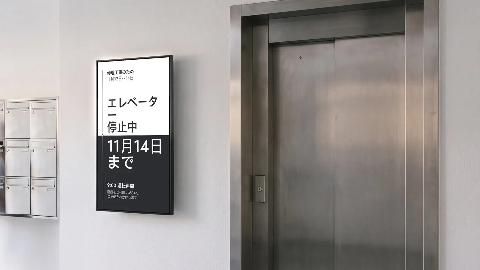マンションのエントランスホール。白い壁のスチール製の集合ポスト(一部のみ写る)と大きなスチール製のエレベーターの扉の間に縦向きの画面がある。画面には、エレベーター停止中、11月14日まで、修理工事は11月12日〜14日、9:00運転再開と表示されている。