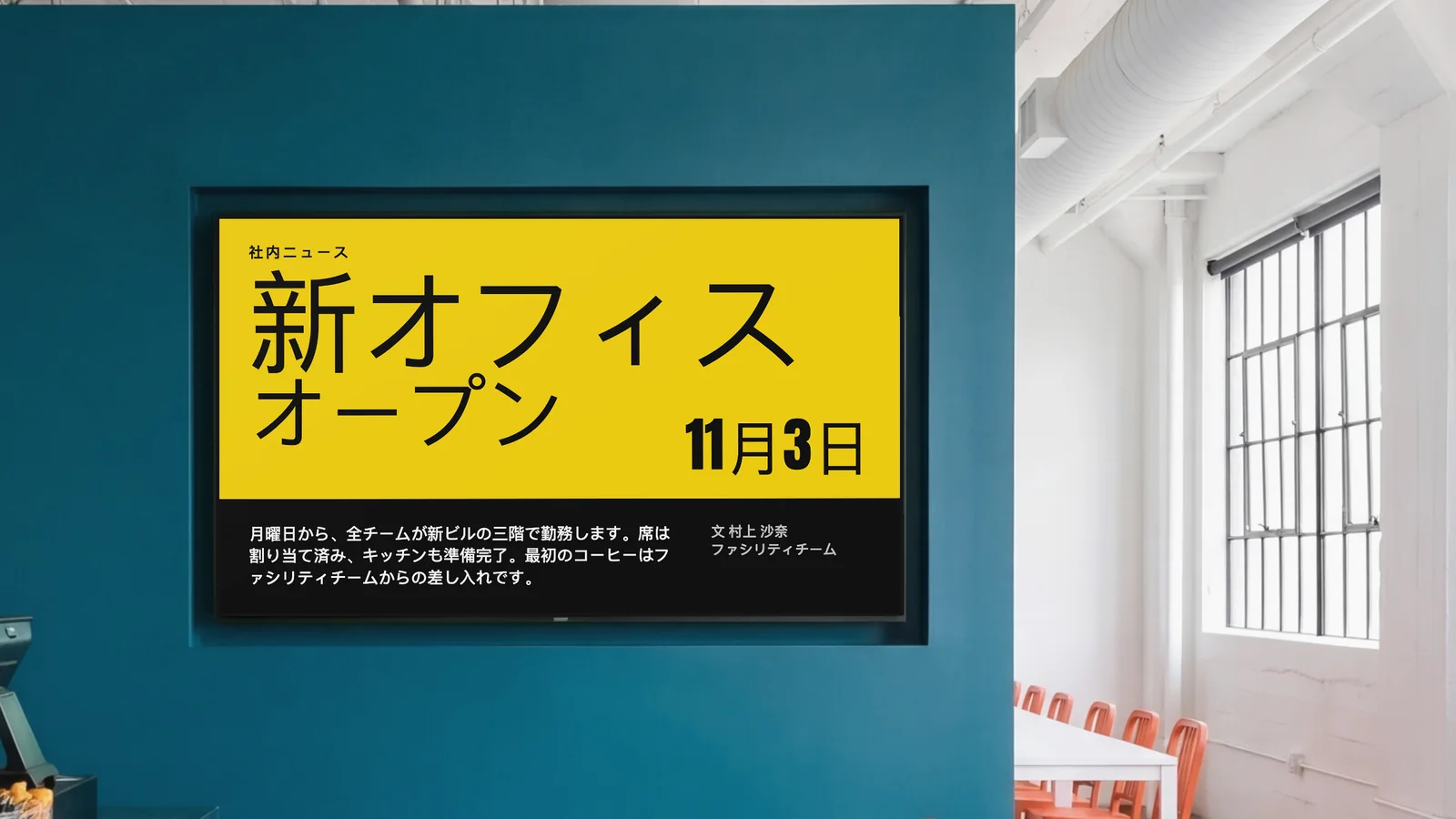 深いティール色の壁に画面が埋め込まれた社員食堂。両脇にカウンターの一部とオレンジ色の椅子が見える。画面には社内ニュースとして、新オフィスが11月3日にオープンすると表示されている。