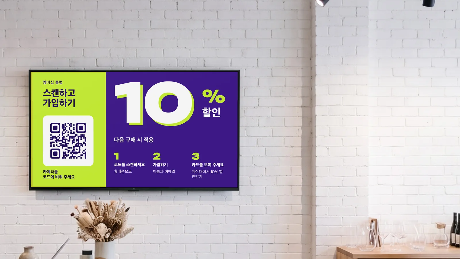 흰 벽돌 벽의 계산대 뒤에 화면이 있는 생활용품 소품샵입니다. 테라조 카운터 위에 드라이플라워가, 옆 선반에 도자기가 놓여 있습니다. 화면에는 멤버십 클럽 안내로 스캔하고 가입하기, 다음 구매 시 10% 할인이 보입니다.