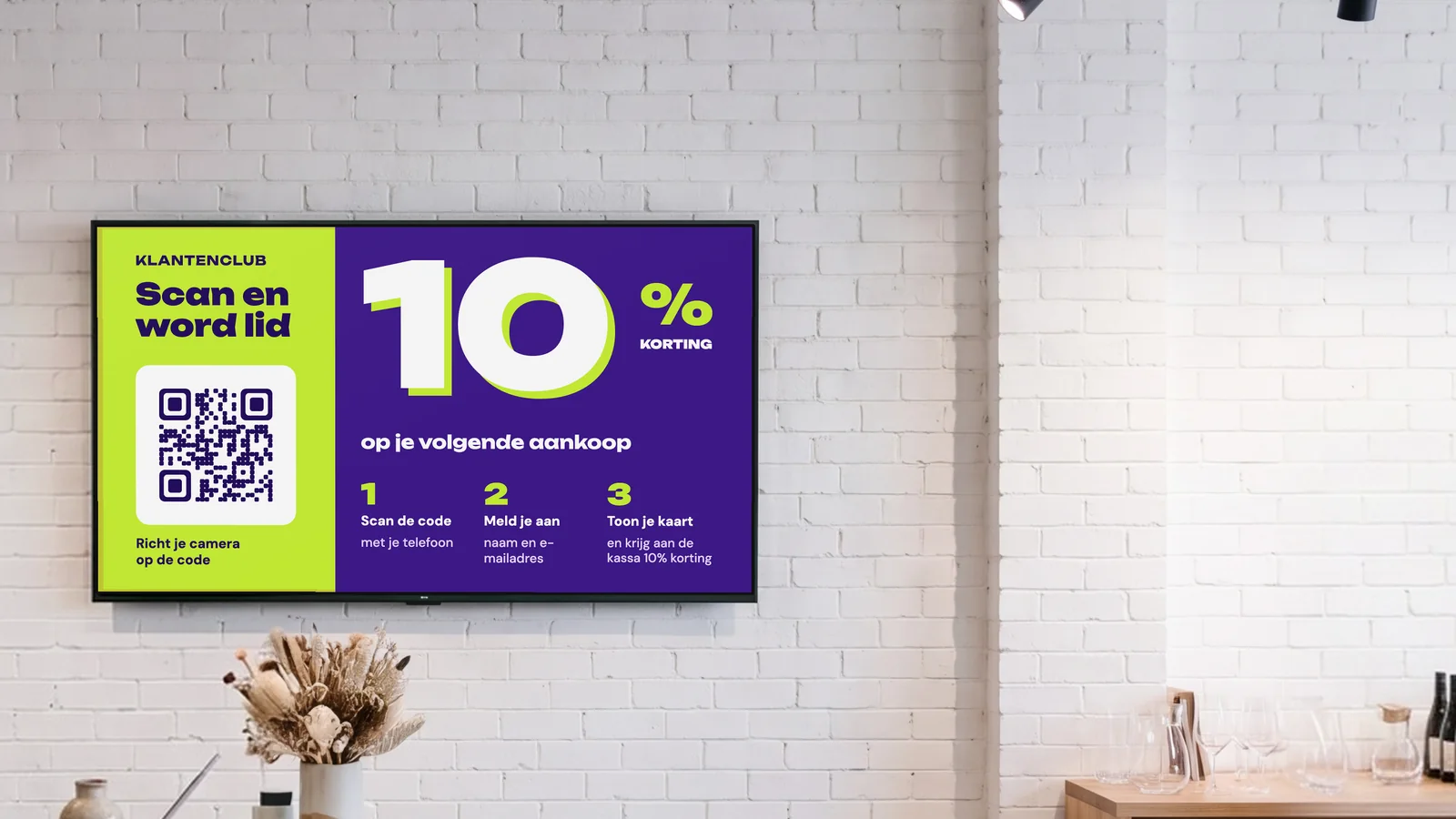 Cadeau- en woonwinkel met een scherm aan de witte bakstenen muur achter de kassa, droogbloemen op een toonbank van terrazzo en keramiek op de schappen ernaast. Het scherm toont de klantenclub: scan en word lid, 10% korting op je volgende aankoop.