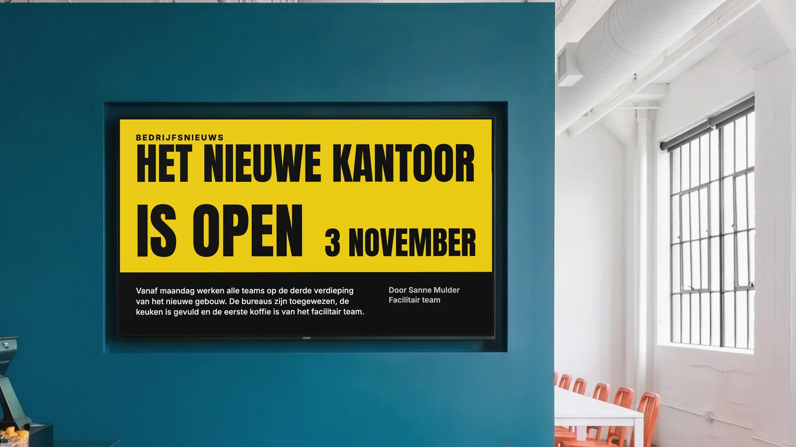Bedrijfskantine met een scherm in een diep petrolkleurige wand, een stukje van de toonbank en oranje stoelen aan de zijkanten. Het scherm meldt bedrijfsnieuws: het nieuwe kantoor is open, 3 november.