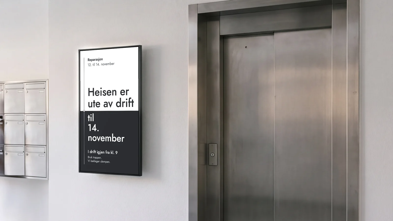 Inngangspartiet i en boligblokk med en stående skjerm på en hvit vegg, mellom postkasser i stål og en stor heisdør i stål. Skjermen viser: Heisen er ute av drift til 14. november, reparasjon 12. til 14. november, i drift igjen fra kl. 9.