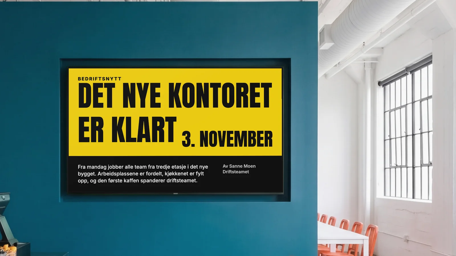 Personalkantine med en skjerm felt inn i en dyp blågrønn vegg, et glimt av disken og oransje stoler på sidene. Skjermen viser Bedriftsnytt: Det nye kontoret er klart 3. november.