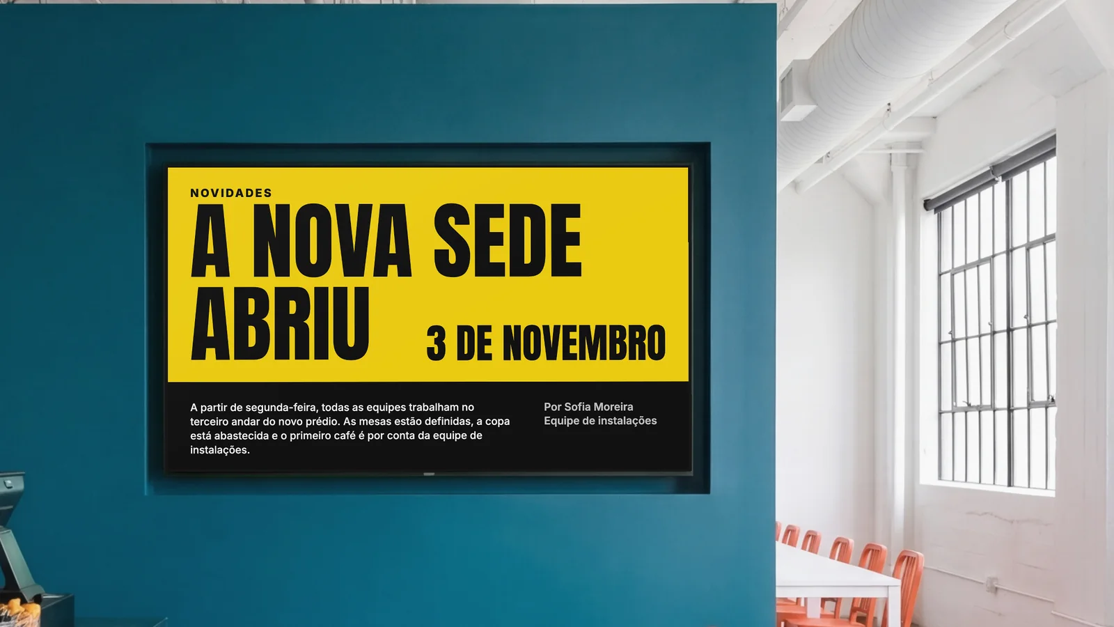 Refeitório de uma empresa com uma tela embutida numa parede azul-petróleo escura, um canto do balcão e cadeiras laranja dos lados. A tela mostra Novidades: a nova sede abriu, 3 de novembro.