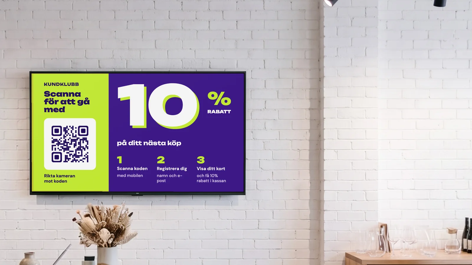 Butik för presenter och inredning med en skärm på den vita tegelväggen bakom kassan, torkade blommor på en disk i terrazzo och keramik på hyllorna bredvid. Skärmen visar kundklubben: scanna för att gå med, 10 % rabatt på ditt nästa köp.