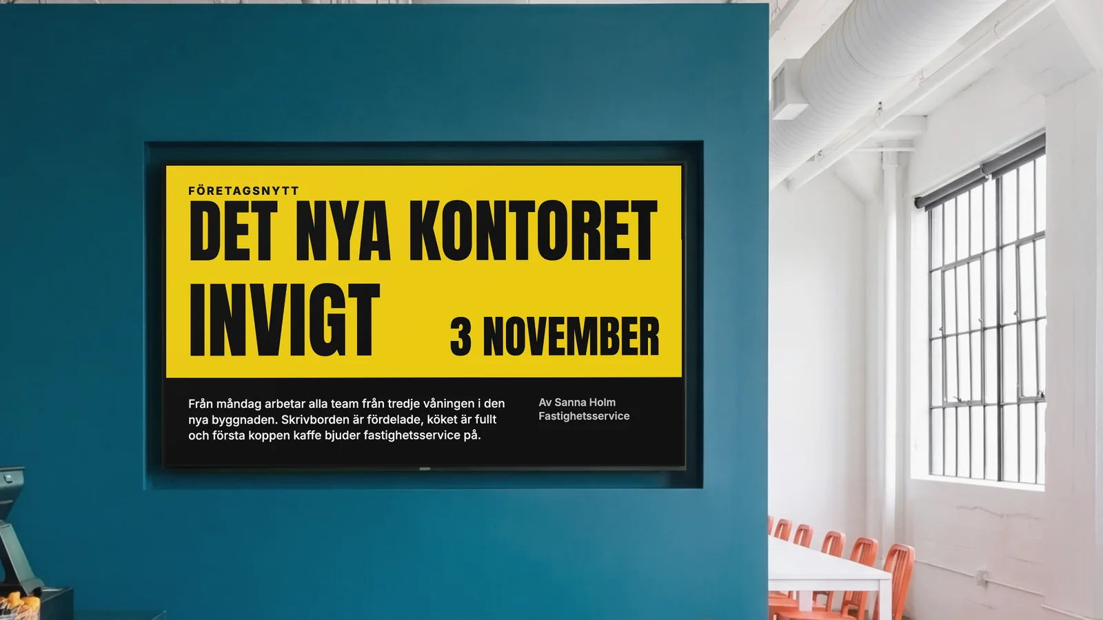 Personalmatsal med en skärm infälld i en djup petrolblå vägg, ett hörn av disken och orange stolar på sidorna. Skärmen visar Företagsnytt: Det nya kontoret invigt 3 november.